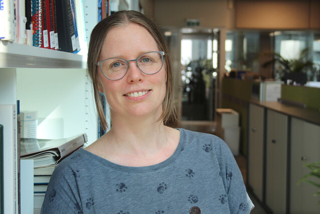 A young blond woman in her thirties with glasses and a blue shirt smiles at the camera. She is standing next to a bookshelf.