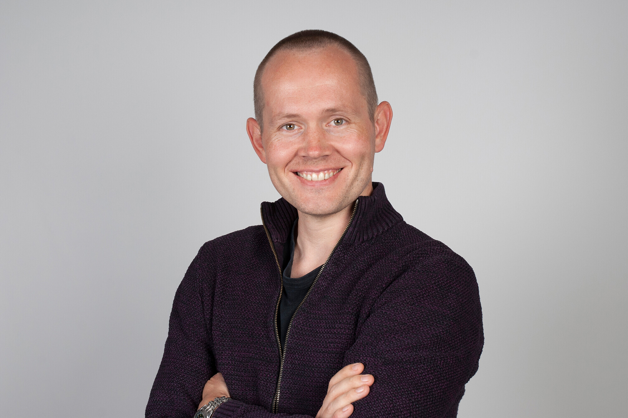 A man is standing and facing the camera with a smile, his arms are crossed and he is wearing a black jacket.