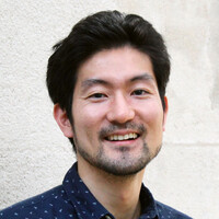 A young Asian man with short black hair is smiling at the camera; he is standing in front of a whitish wall.