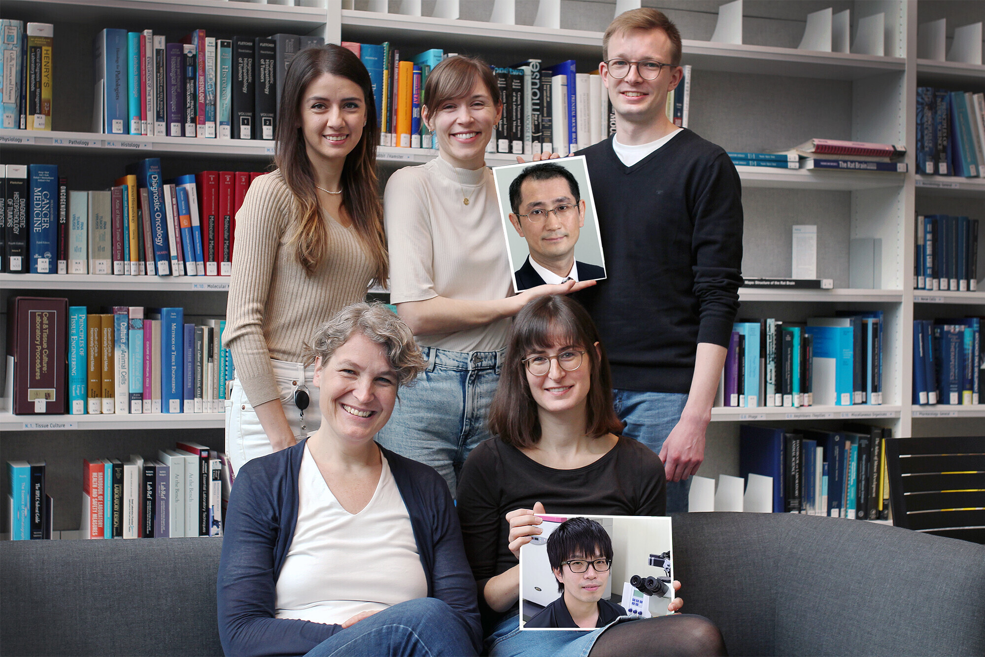 A group of five people smiles at the camera, 4 women one man. Two of the women hold frames displaying the faces of two other men.