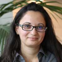 A young women with dark glasses and long dark hair smiles at the camera; she is standing in front of a plant.