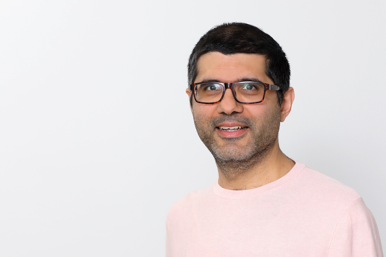 A man with short black hair and brown glasses is smiling to the camera. He wears a light pink sweater.