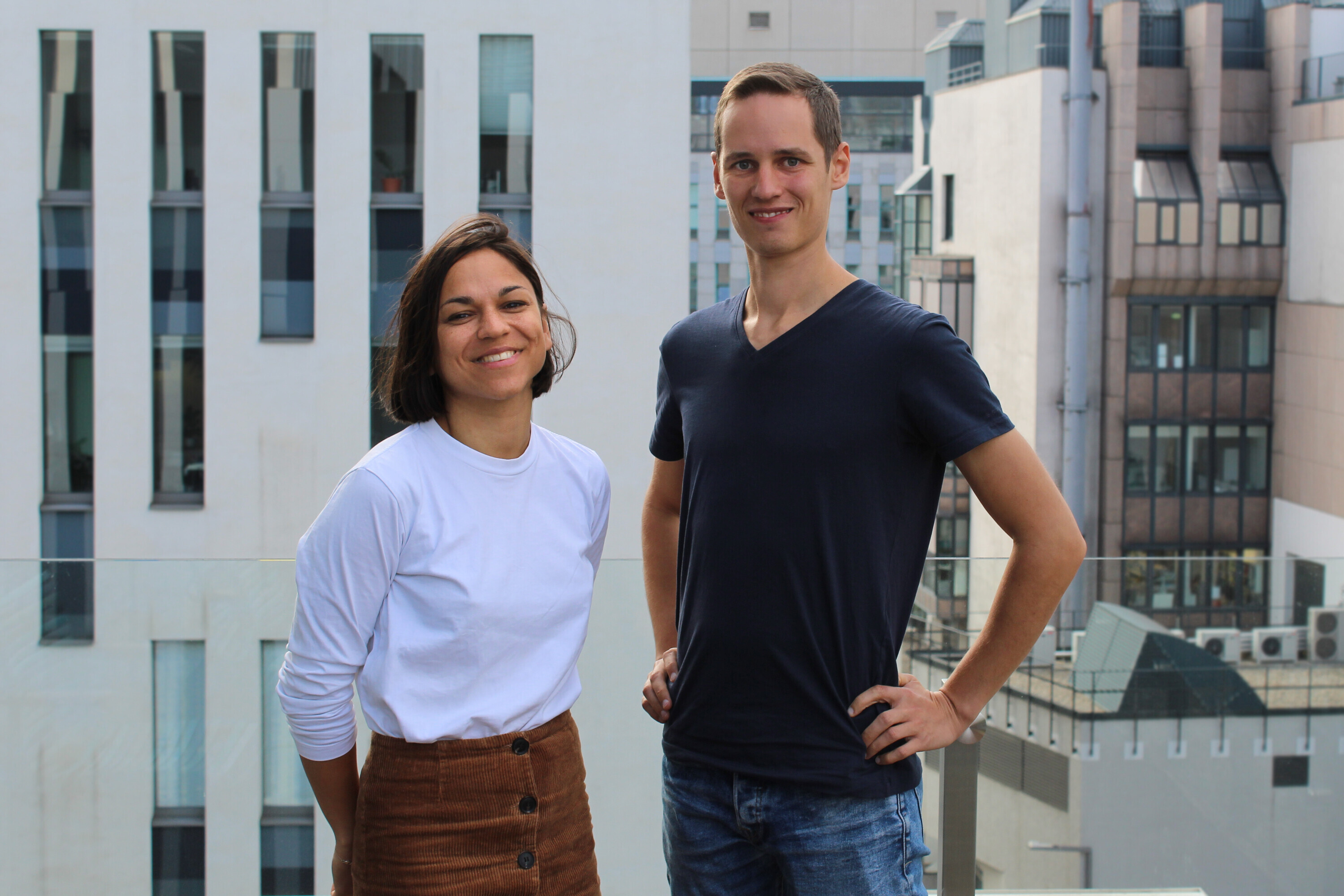 Two young people are standing on a terrace. On the left, a woman with a white shirt and a brown skirt, with short black hair, is smiling. On the right, a tall man with a dark shirt and jeans is smiling.