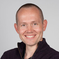 A young white man with buzzed hair smiles at the camera. He is wearing a dark sweater and stands in front of a grey background.