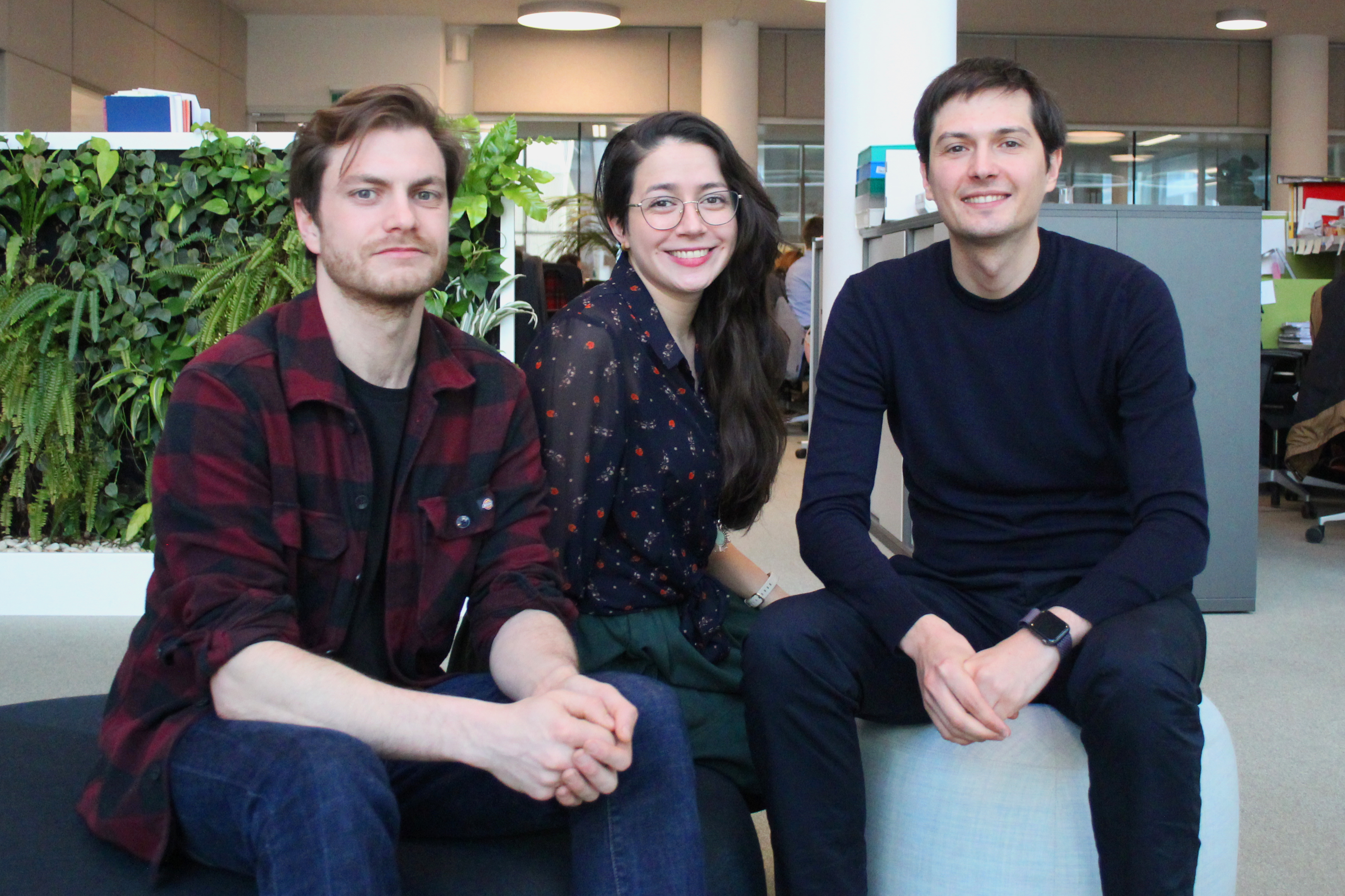 Three young people sit and smile at the camera. From left to right: a man with brown hair, a red shirt and jeans; a woman with long dark hair, glasses, a dark blue top, and a dark green skirt; and a man with brown hair, a dark blue top and jeans.