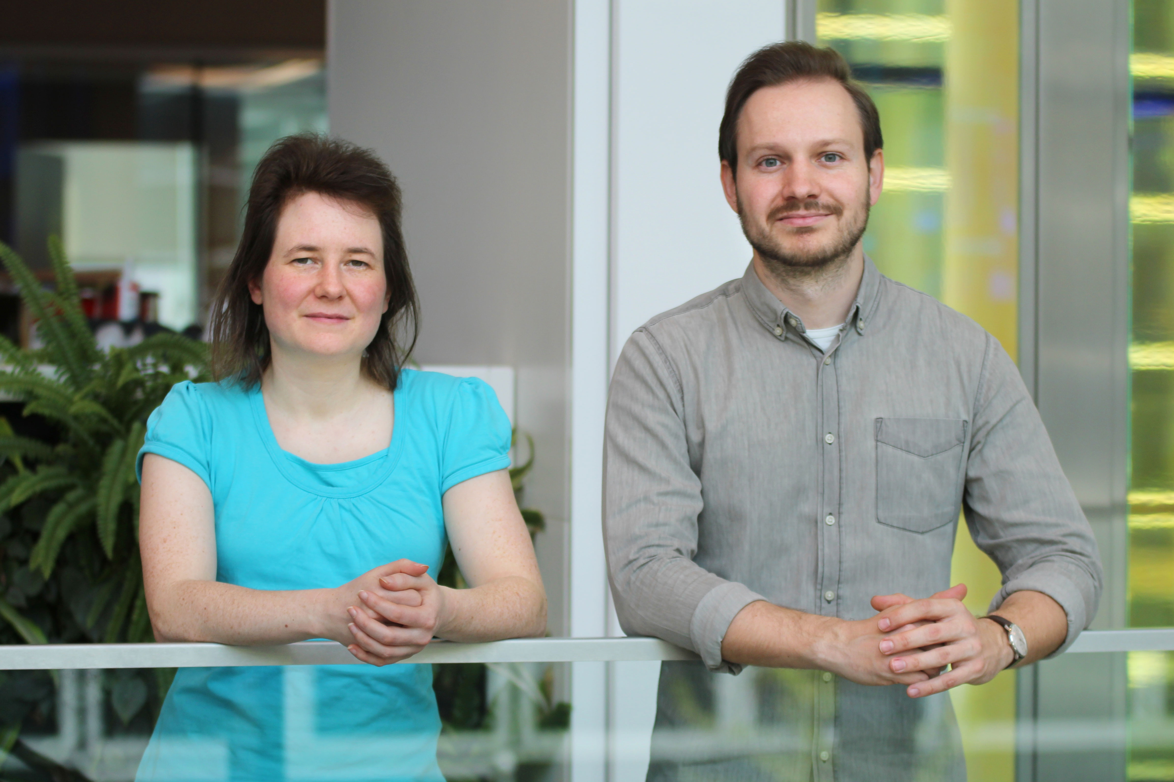 A young woman dressed in turquoise and a young man with a grey shirt lean on a glass wall and look at the camera.