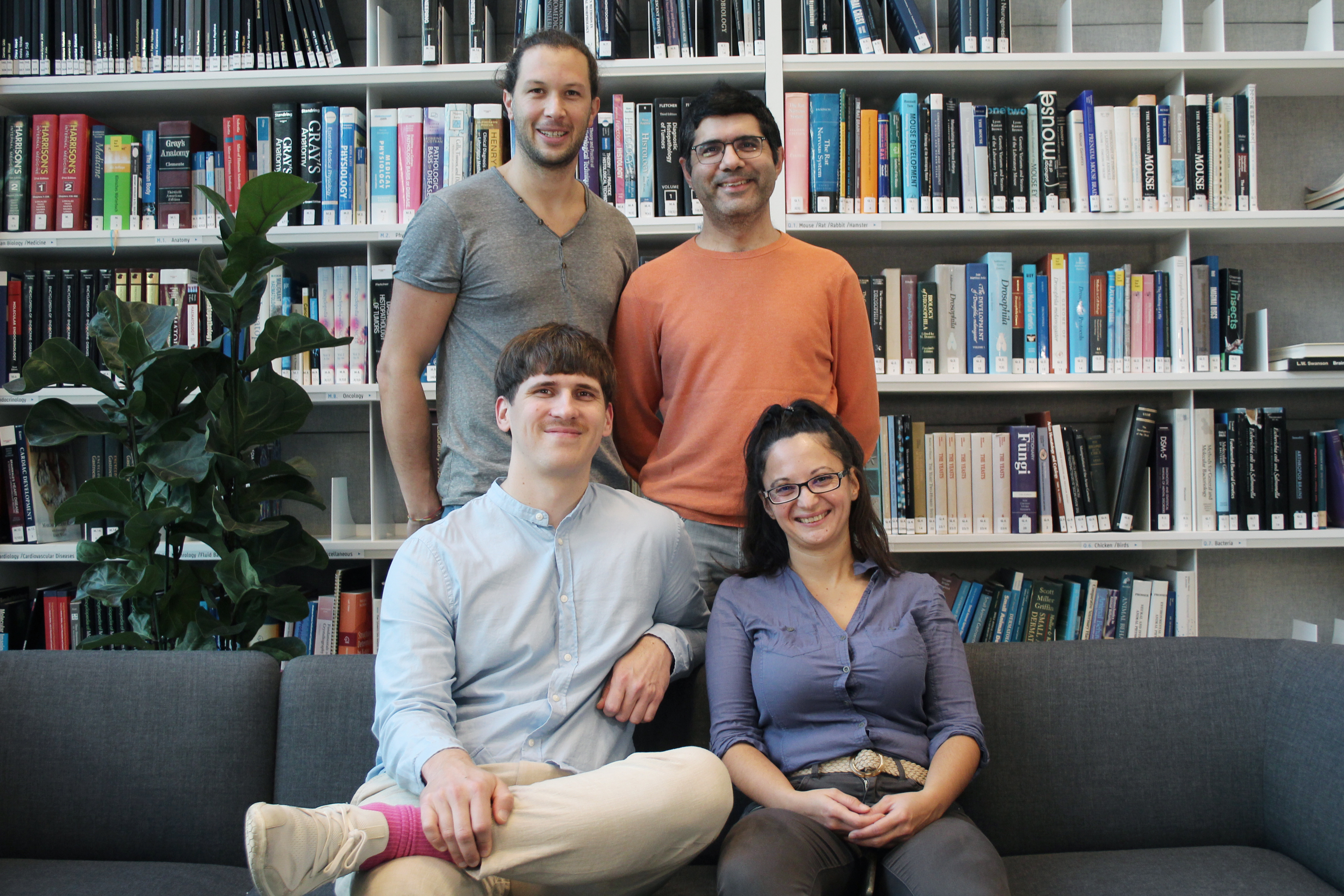 Four people in two rows, two sitting on a couch and two standing behind them, smiling at the camera. Behind them is a massive bookshelf.